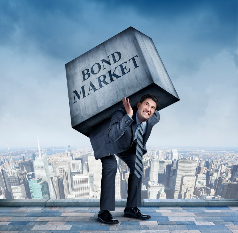 A businessman struggles carries a large concrete block representing the bond market on his back with a large urban skyline in the background.  The image represents the increase in interest rates which results in a falling prices in the bond markets.